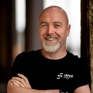 A smiling David McFall wears a black t-shirt with the SAFER GROUP logo while standing confidently with arms crossed, leaning against a stone pillar.