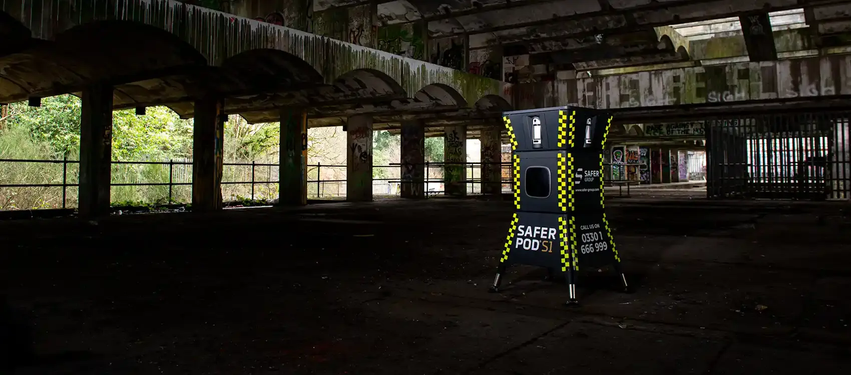 A yellow and black safer POD s1 device stands inside an abandoned, dilapidated building with graffiti and overgrown foliage visible outside.