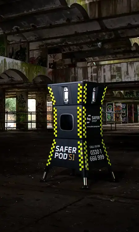 A yellow and black safer POD s1 device stands inside an abandoned, dilapidated building with graffiti and overgrown foliage visible outside.