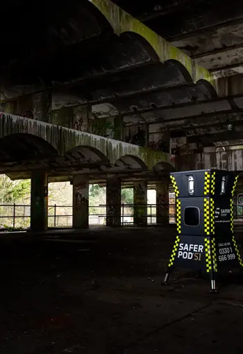 A yellow and black safer POD s1 device stands inside an abandoned, dilapidated building with graffiti and overgrown foliage visible outside.