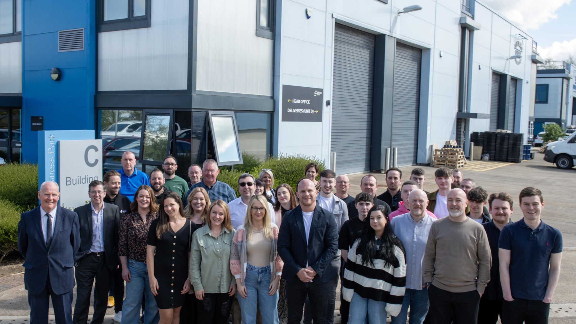 A group photo of employees and council representatives in front of the Safer Group building, showcasing diverse outfits and professional attire.