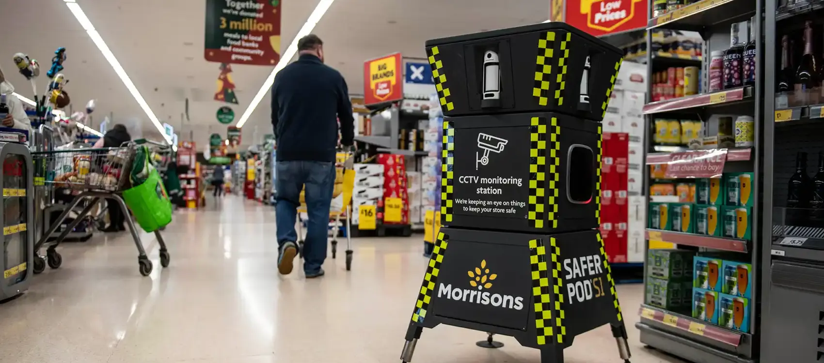 A Safer POD S1 device stands at the end of a grocery store aisle, with shoppers and products visible, promoting safety and surveillance by the supermarket Morrisons.