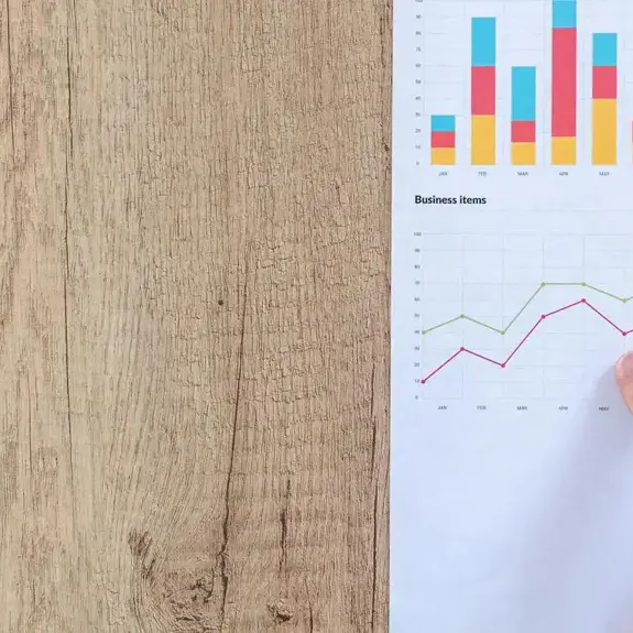 a hand points to a graph trending upwards on a sheet of paper. the paper is resting on a light wooden desk. 