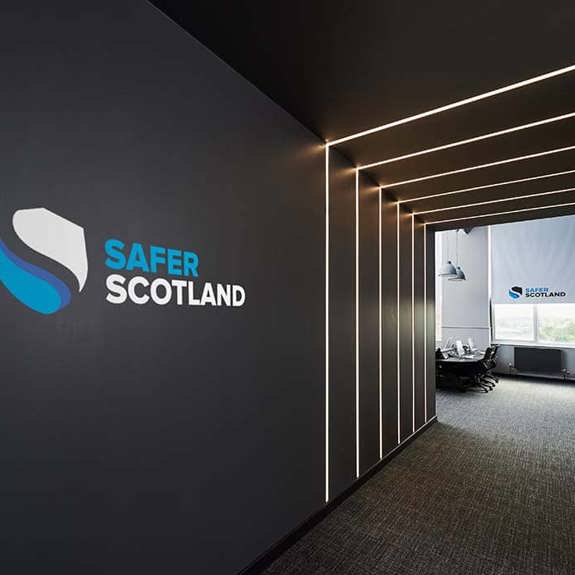A sleek office hallway featuring a "Safer Scotland" logo on the wall, with modern lighting and glass-panelled meeting rooms.