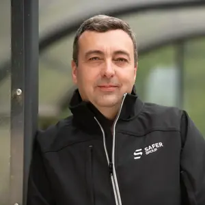 Phil Bunting stands wearing a black zip up jacket with the "SAFER GROUP" logo outdoors beside a glass structure, greenery in the background.