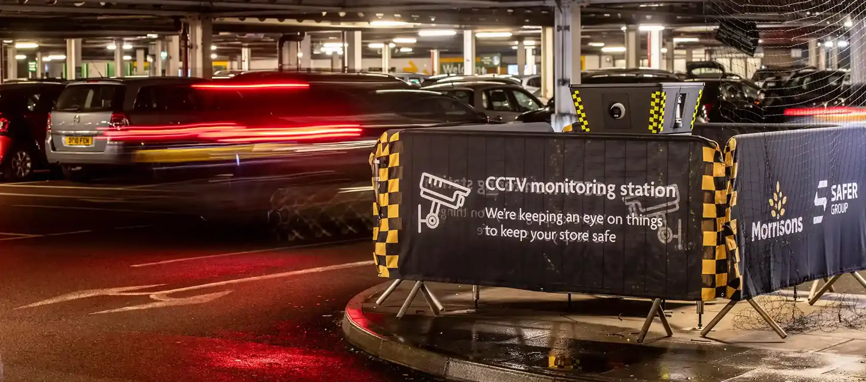 A dimly lit parking garage features a Safer POD S4 CCTV monitoring station surrounded by a morrisons banner, while a blurred car speeds past.