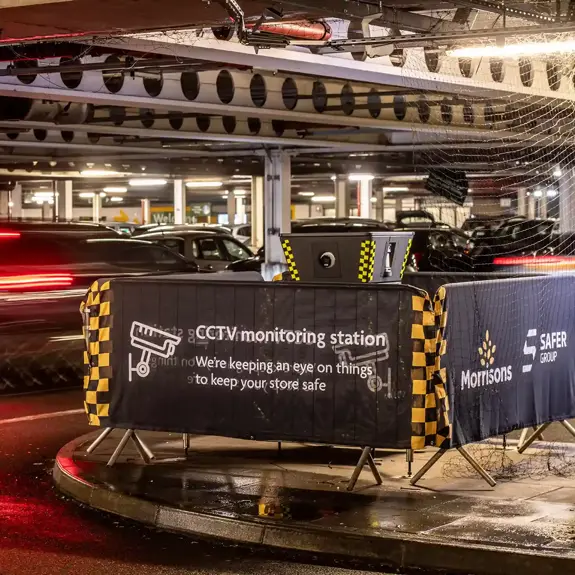 A dimly lit parking garage features a Safer POD S4 CCTV monitoring station surrounded by a morrisons banner, while a blurred car speeds past.