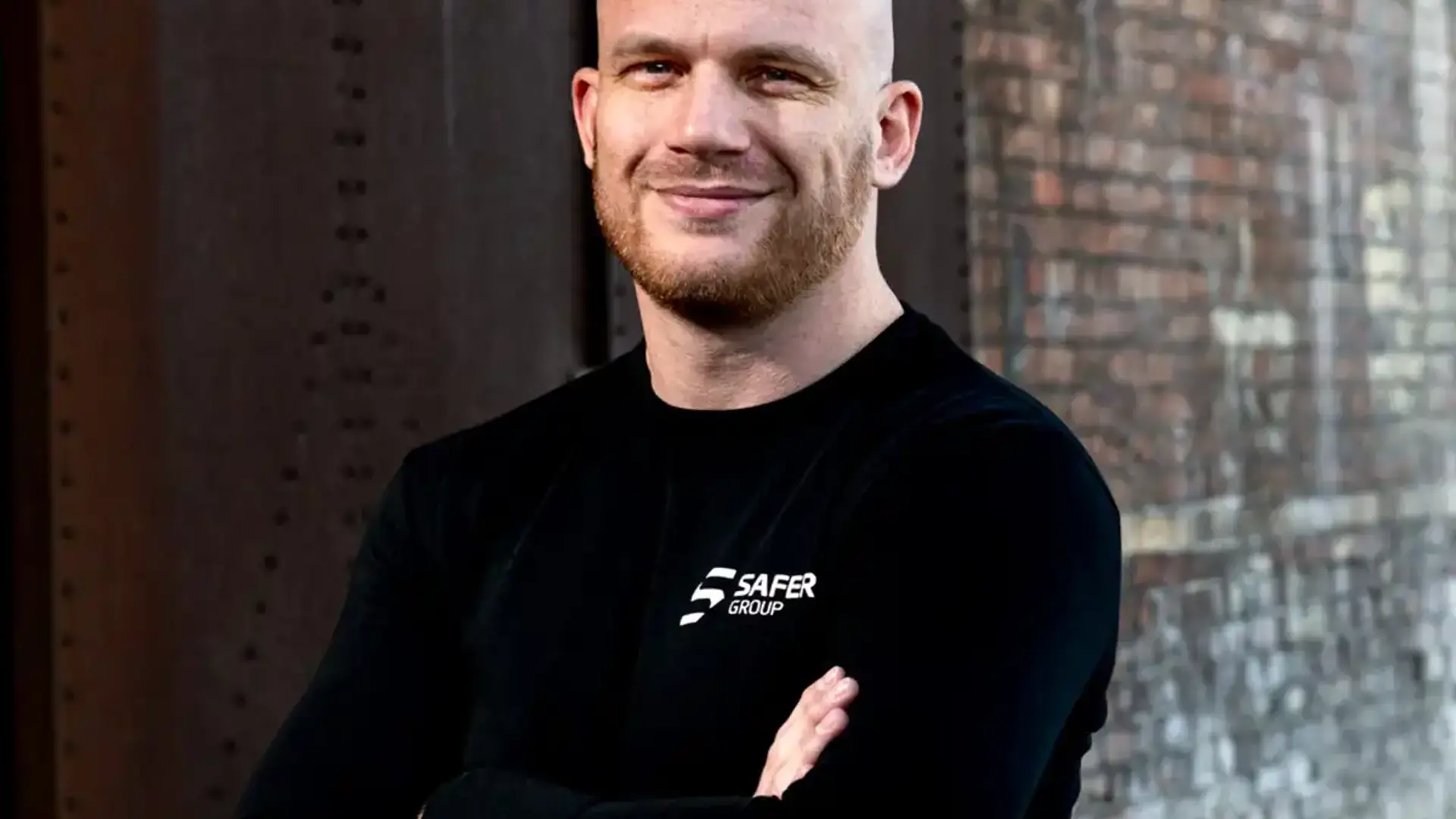Ryan Clark stands confidently in a black long-sleeve shirt with arms crossed against a textured brick wall.