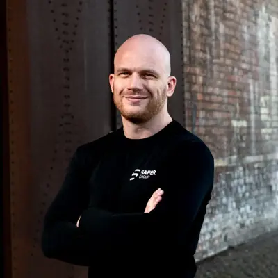 Ryan Clark stands confidently in a black long-sleeve shirt with arms crossed against a textured brick wall.