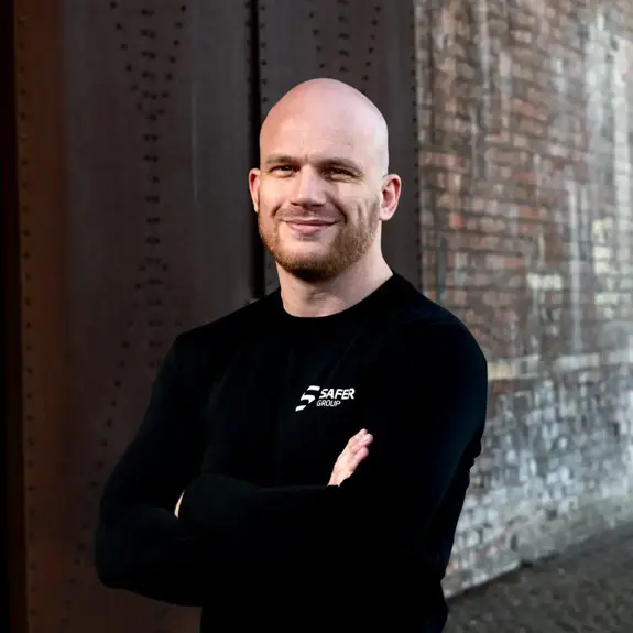 Ryan Clark stands confidently in a black long-sleeve shirt with arms crossed against a textured brick wall.