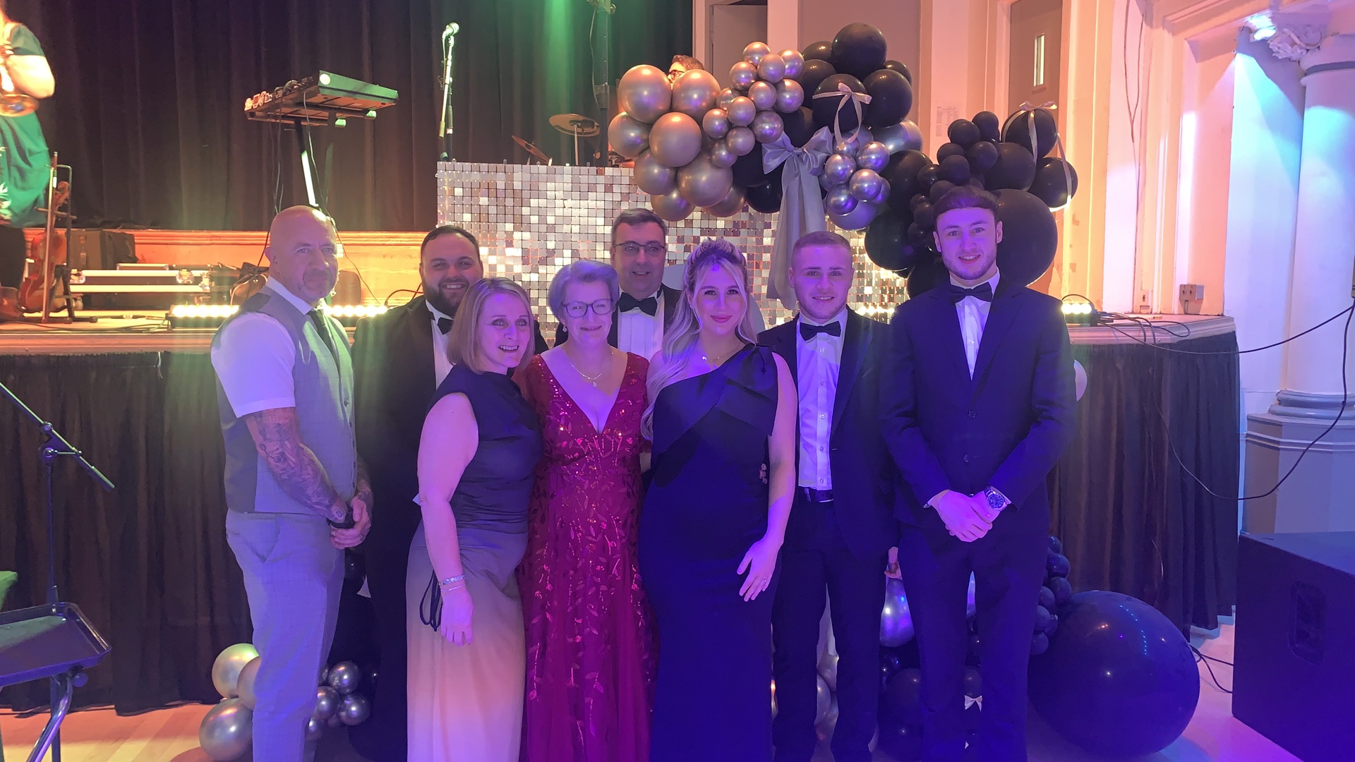 A group of elegantly dressed individuals pose in front of a decorated stage with balloons, lights, and musical instruments visible.