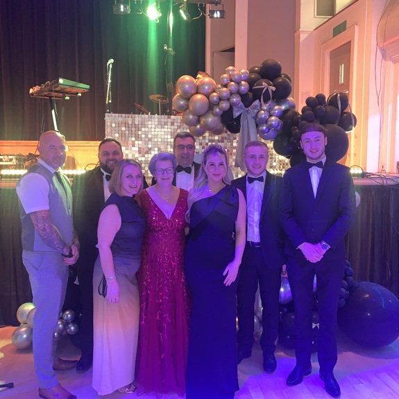 A group of elegantly dressed individuals pose in front of a decorated stage with balloons, lights, and musical instruments visible.