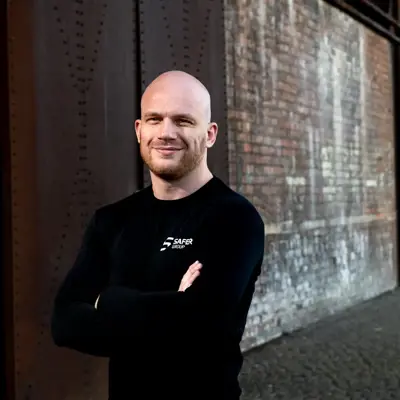 Ryan Clark stands confidently in a black long-sleeve shirt with arms crossed in front of a textured brick wall.