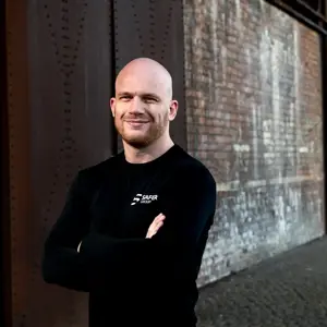 Ryan Clark stands confidently in a black long-sleeve shirt with arms crossed in front of a textured brick wall.