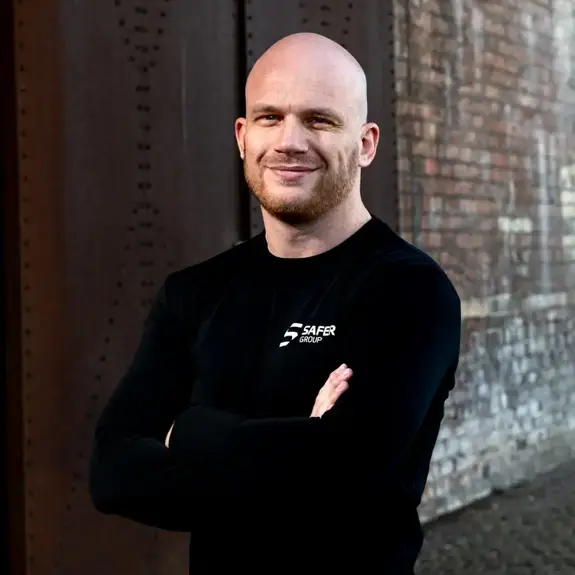 Ryan Clark stands confidently in a black long-sleeve shirt with arms crossed in front of a textured brick wall.