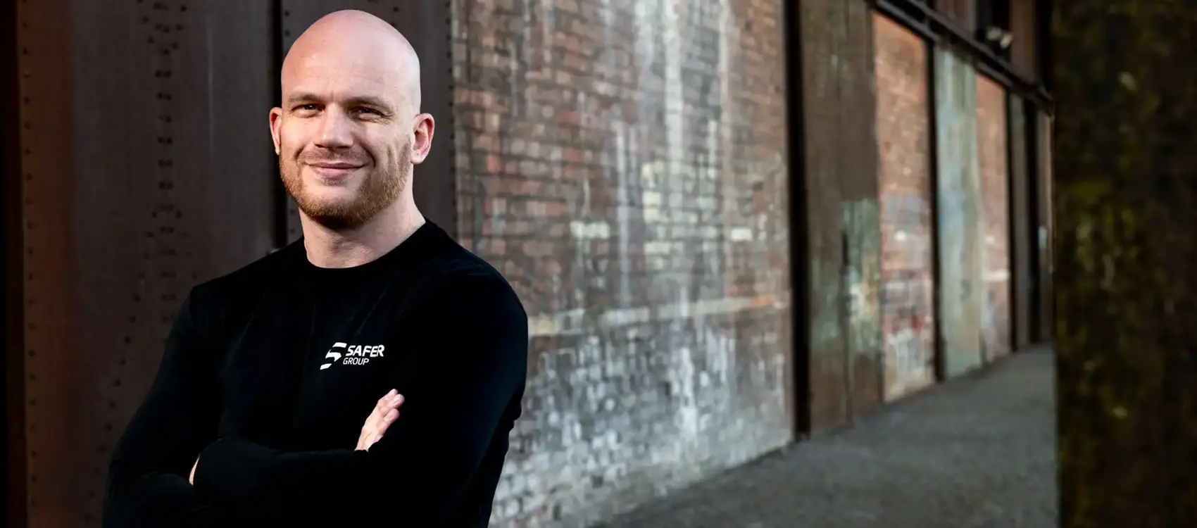 Ryan Clark stands confidently in a black long-sleeve shirt with arms crossed in front of a textured brick wall.