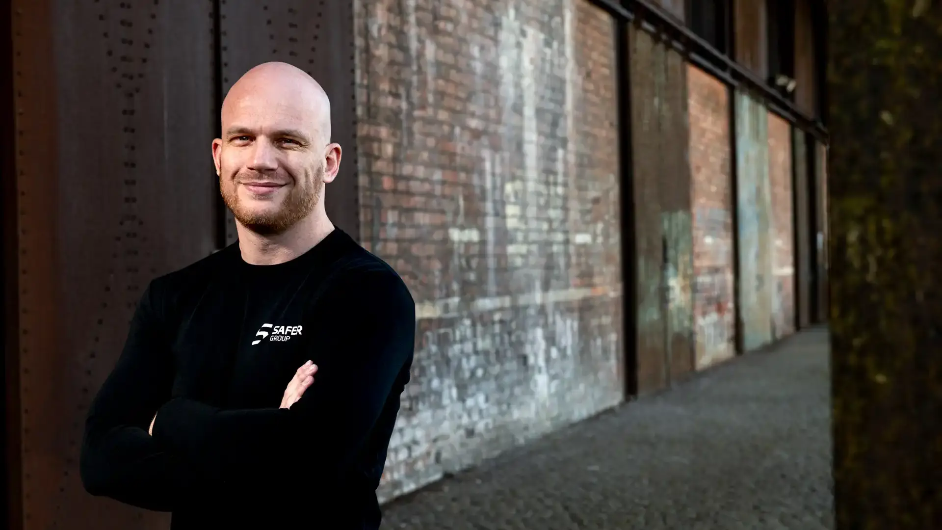 Ryan Clark stands confidently in a black long-sleeve shirt with arms crossed in front of a textured brick wall.