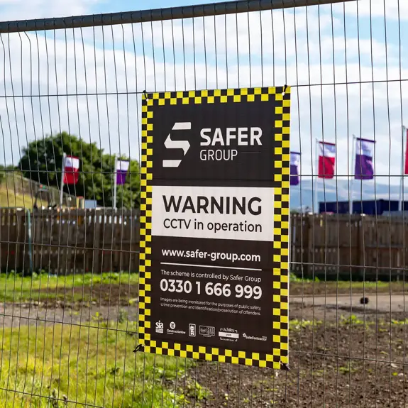 A sign on a construction site fence warns of CCTV in operation by Safer Group, set against a background of colourful flags and buildings.