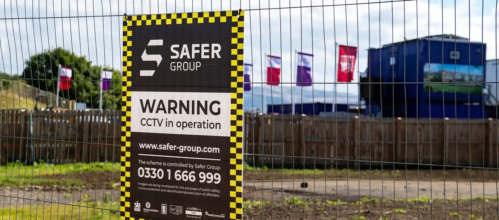 A sign on a construction site fence warns of CCTV in operation by Safer Group, set against a background of colourful flags and buildings.