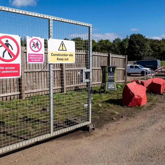 A gated construction site with warning signs stating No access for pedestrians and Children not allowed to play. In the background, a safer POD S1 sits behind two large red stones providing security.