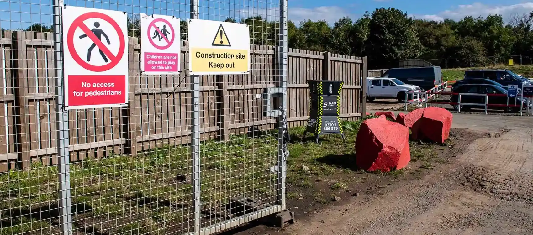 A gated construction site with warning signs stating No access for pedestrians and Children not allowed to play. In the background, a safer POD S1 sits behind two large red stones providing security.