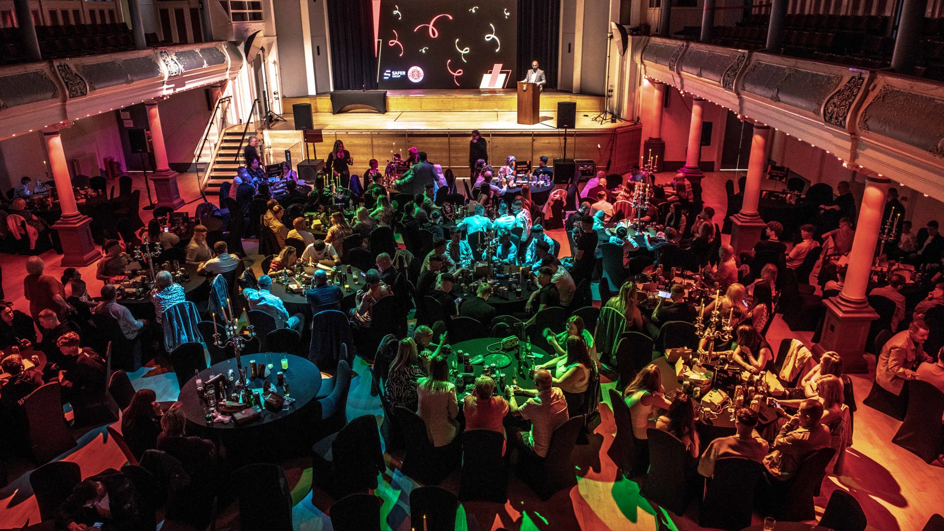 A formal gathering in a grand hall with round tables filled with people under colourful lights. A stage with a speaker and large screen is in the background.