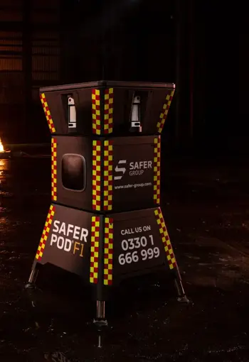 Two black safer pod F1s with yellow and red reflective patterns stand in a dimly lit space, flames flicker in the background alongside a Safer POD F7.