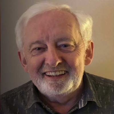 A man with grey hair and trimmed beard wears a patterned shirt, seated in a softly lit room. The background is slightly blurred.