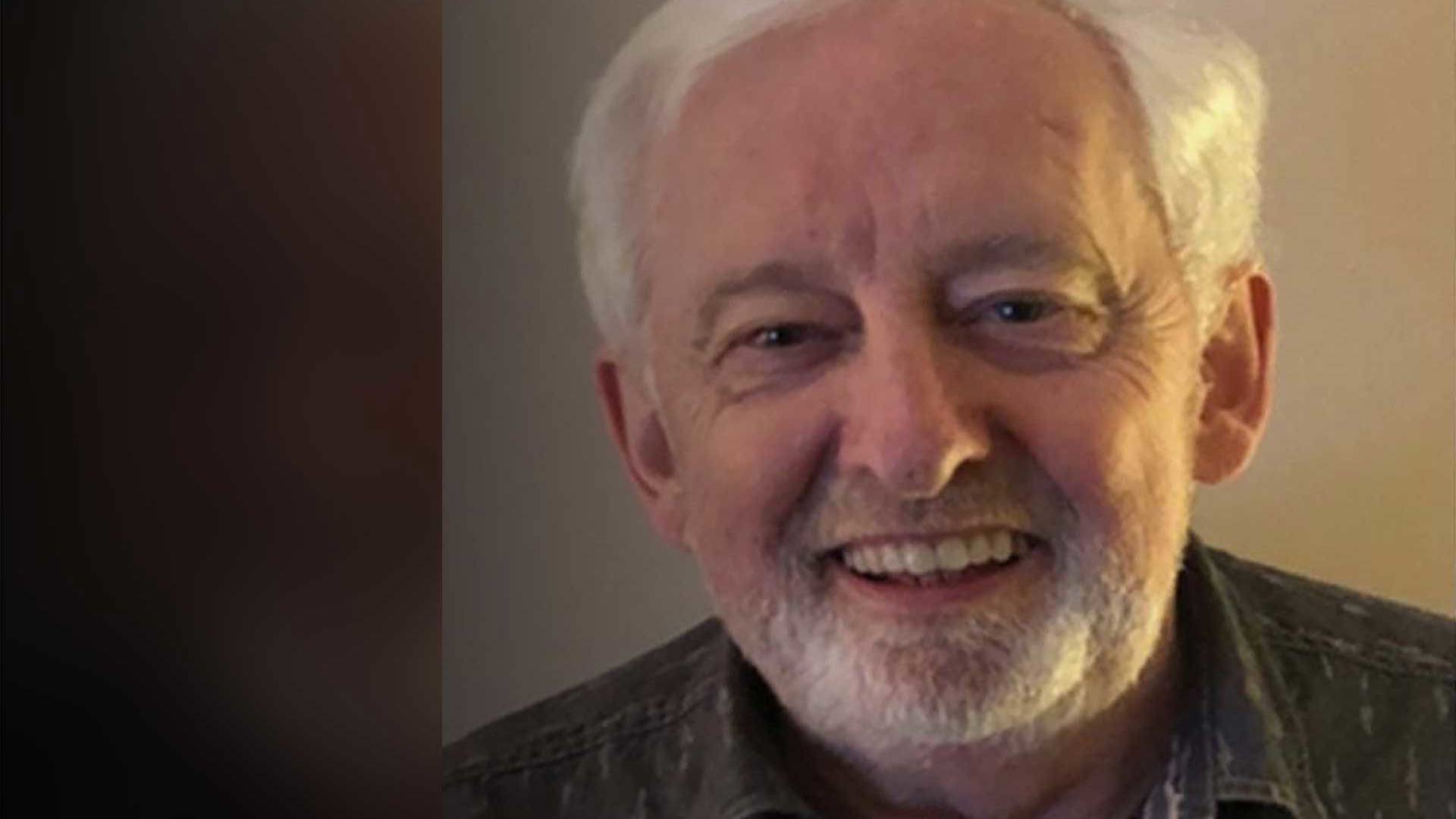 A man with grey hair and trimmed beard wears a patterned shirt, seated in a softly lit room. The background is slightly blurred.