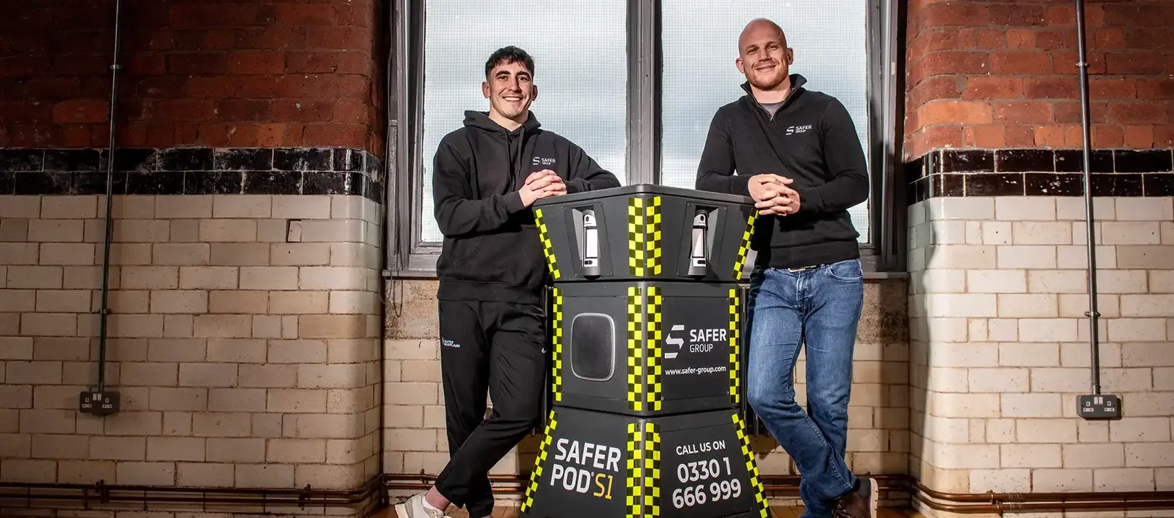 MMA fighter Reece McEwan and Group chief executive Ryan Clark stand leaning on either side of a Safer Pod S1, in a rustic brick-walled room in front of large windows.