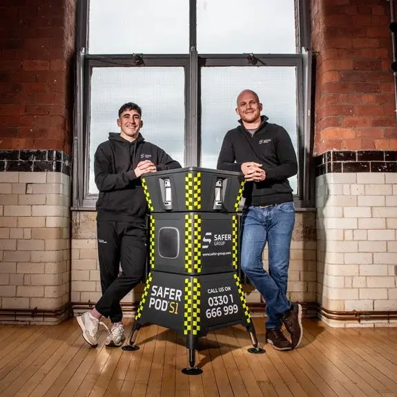 MMA fighter Reece McEwan and Group chief executive Ryan Clark stand leaning on either side of a Safer Pod S1, in a rustic brick-walled room in front of large windows.