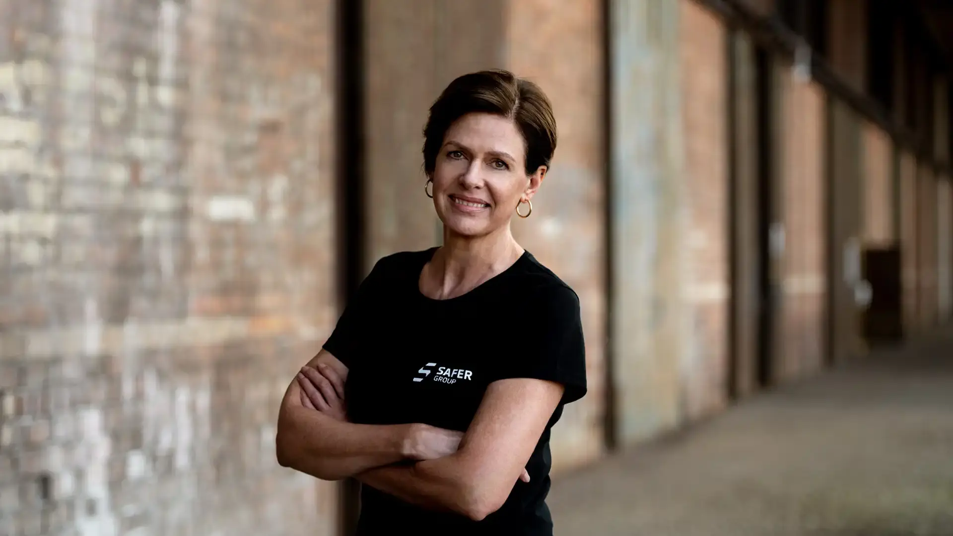 Jan Brazier stands confidently with arms crossed, wearing a black t-shirt with "SAFER GROUP" logo, set against a rustic brick background.