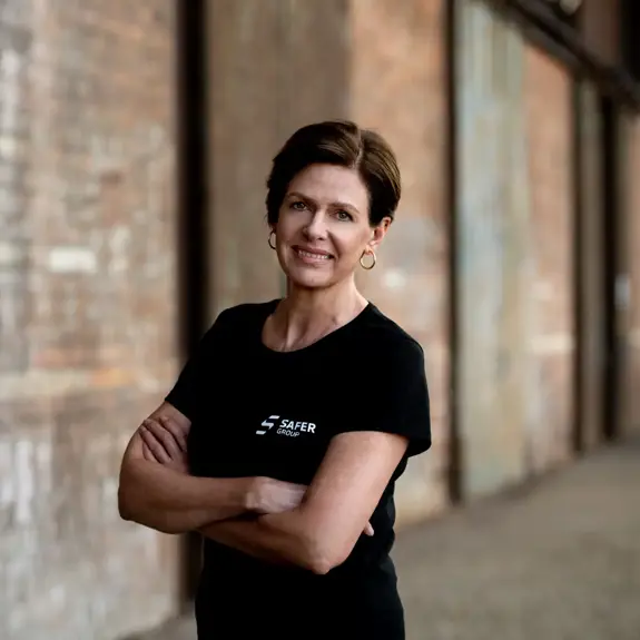 Jan Brazier stands confidently with arms crossed, wearing a black t-shirt with "SAFER GROUP" logo, set against a rustic brick background.