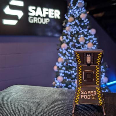 A small toy of a Safer Pod S1 device stands in the foreground, with a Safer Group logo backdrop and a decorated Christmas tree adorned with white lights.