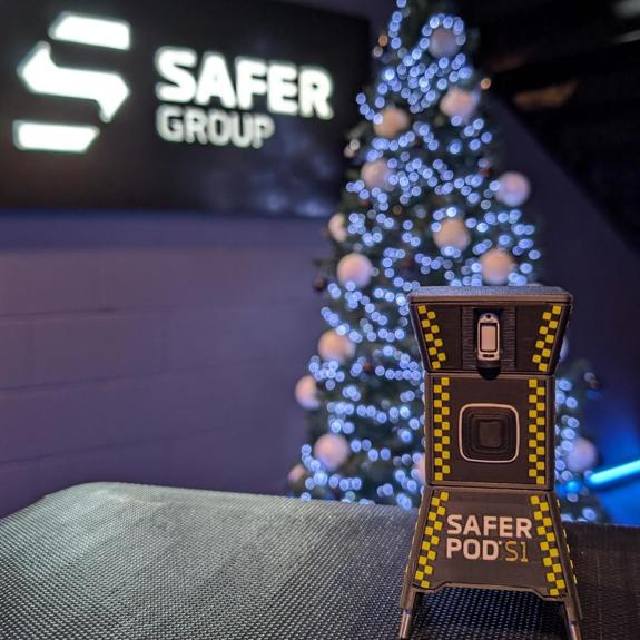 A small toy of a Safer Pod S1 device stands in the foreground, with a Safer Group logo backdrop and a decorated Christmas tree adorned with white lights.