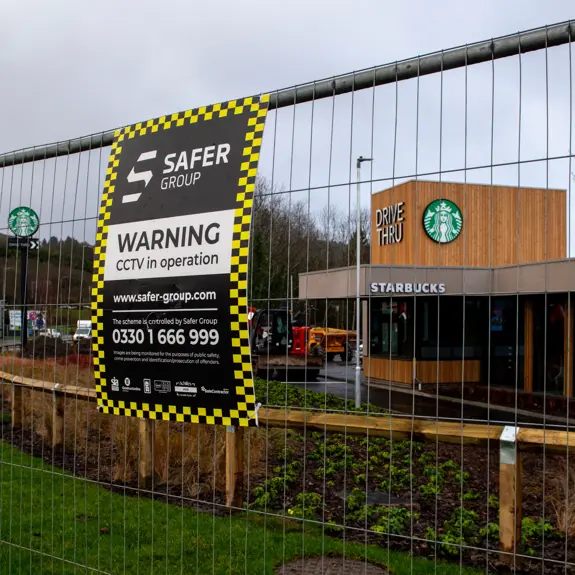 A Starbucks drive-thru location, partially hidden by construction fencing featuring a Safer Group CCTV warning sign.