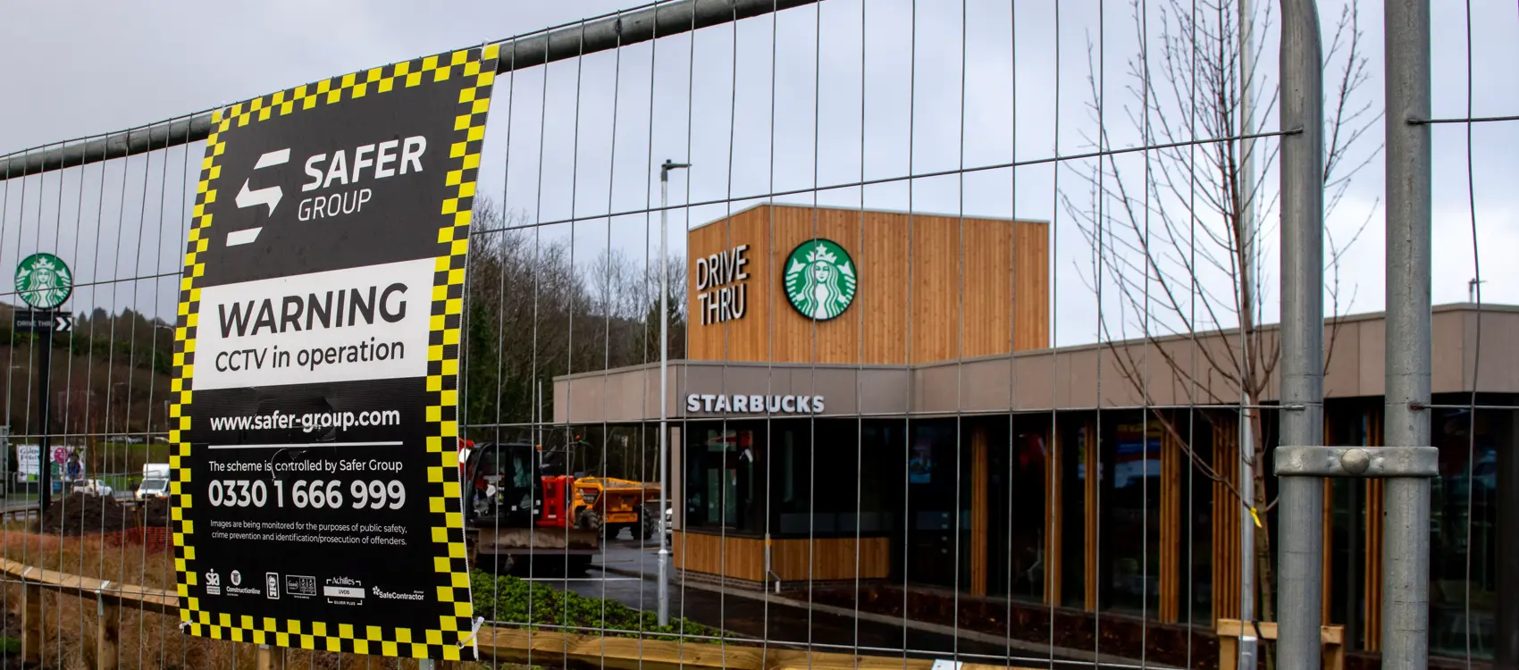 A Starbucks drive-thru location, partially hidden by construction fencing featuring a Safer Group CCTV warning sign.