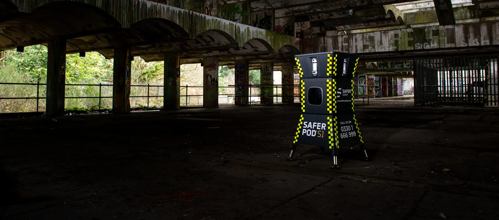 A yellow and black security pod stands inside an abandoned, dilapidated building with graffiti and overgrown foliage visible outside.
