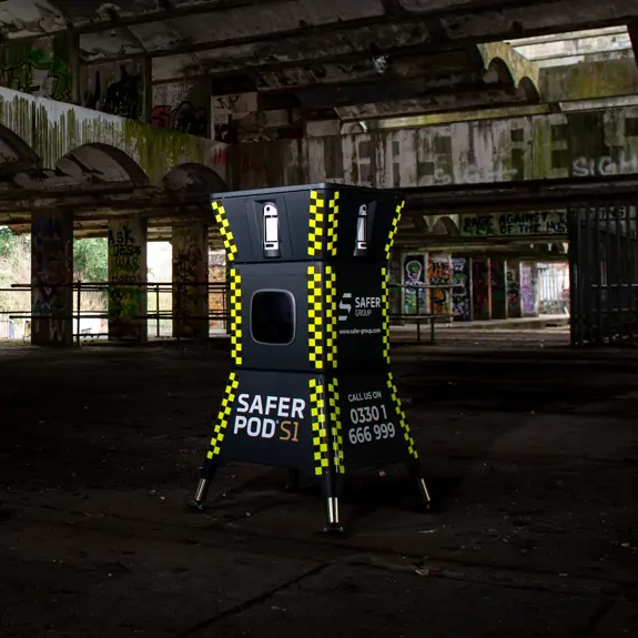 A yellow and black security pod stands inside an abandoned, dilapidated building with graffiti and overgrown foliage visible outside.
