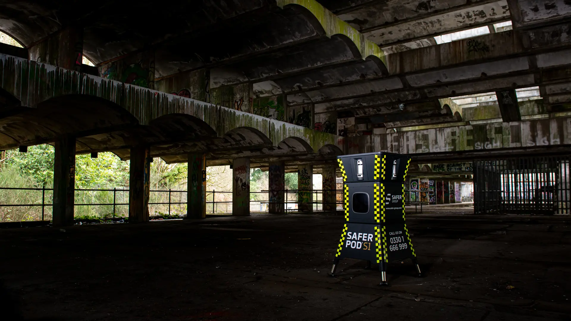 A yellow and black security pod stands inside an abandoned, dilapidated building with graffiti and overgrown foliage visible outside.