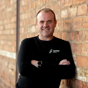 Keith Miller leans against a rustic brick wall, arms folded, in a black long-sleeve shirt with the safer group logo.