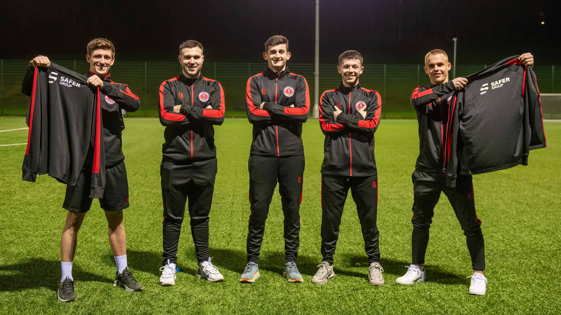 Five footballers in matching black and red tracksuits stand confidently on a sports field, showcasing jackets with the safer group brand.