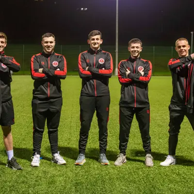 Five footballers in matching black and red tracksuits stand confidently on a sports field, showcasing jackets with the safer group brand.