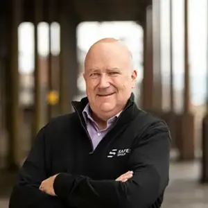 Alastair Russell stands confidently with arms crossed, wearing a black jacket with a Safer Group logo, against a blurred industrial background.