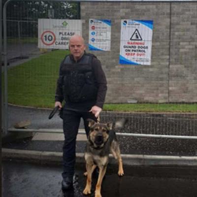 A security officer in tactical gear stands with a German Shepherd outside a gated area. Safer Scotland warning signs are visible nearby.