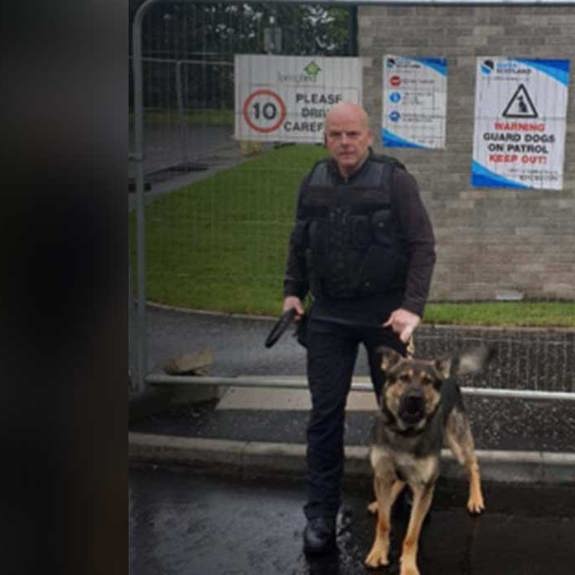 A security officer in tactical gear stands with a German Shepherd outside a gated area. Safer Scotland warning signs are visible nearby.