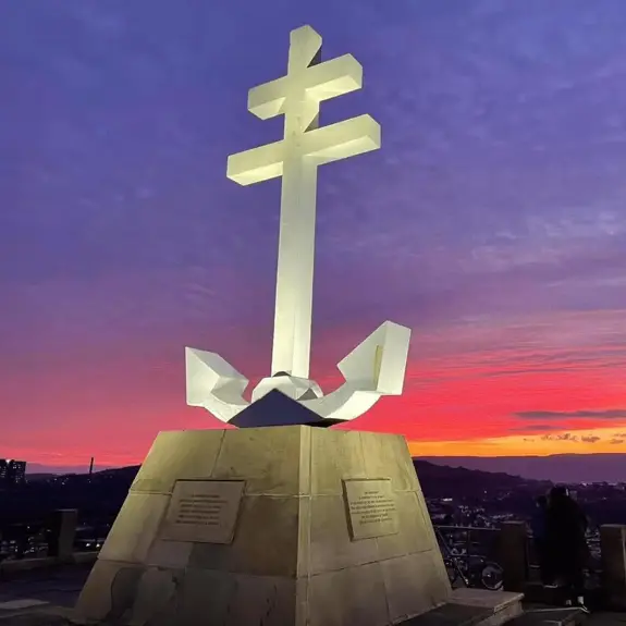 A large white cross atop a monument with an anchor, set against a vibrant sunset sky of purples and oranges.