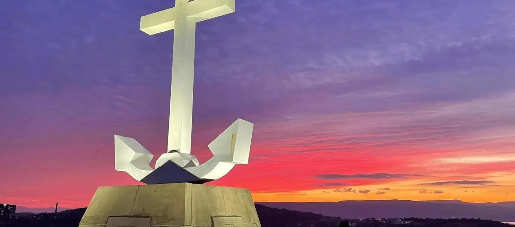 A large white cross atop a monument with an anchor, set against a vibrant sunset sky of purples and oranges.