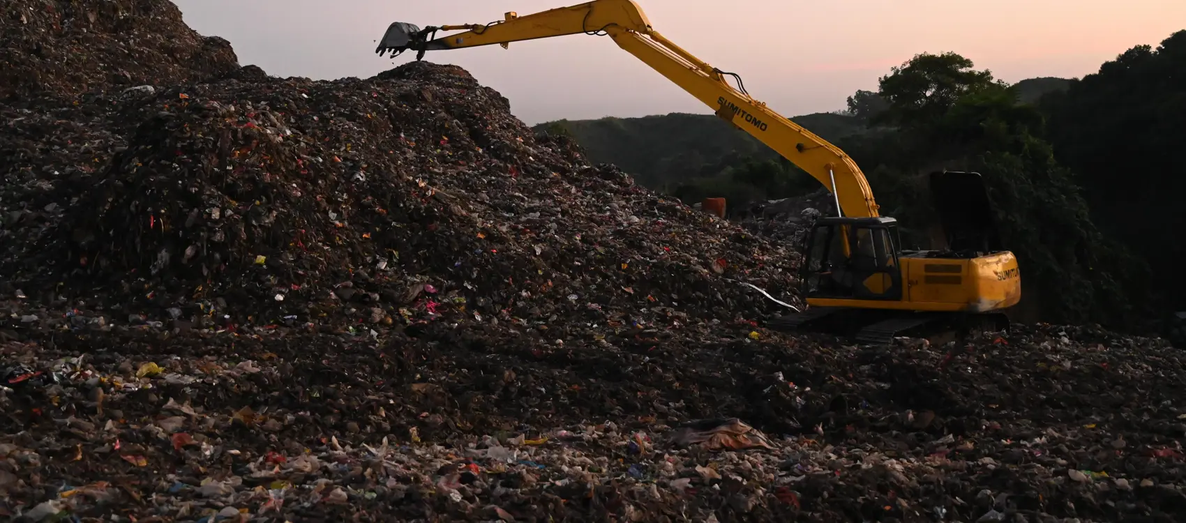 A yellow excavator moves debris atop a large garbage mound at sunset, highlighting the scale of waste management efforts.
