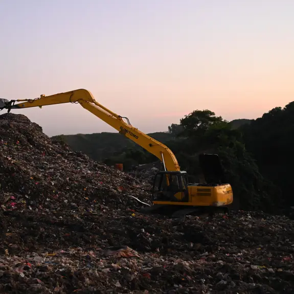 A yellow excavator moves debris atop a large garbage mound at sunset, highlighting the scale of waste management efforts.